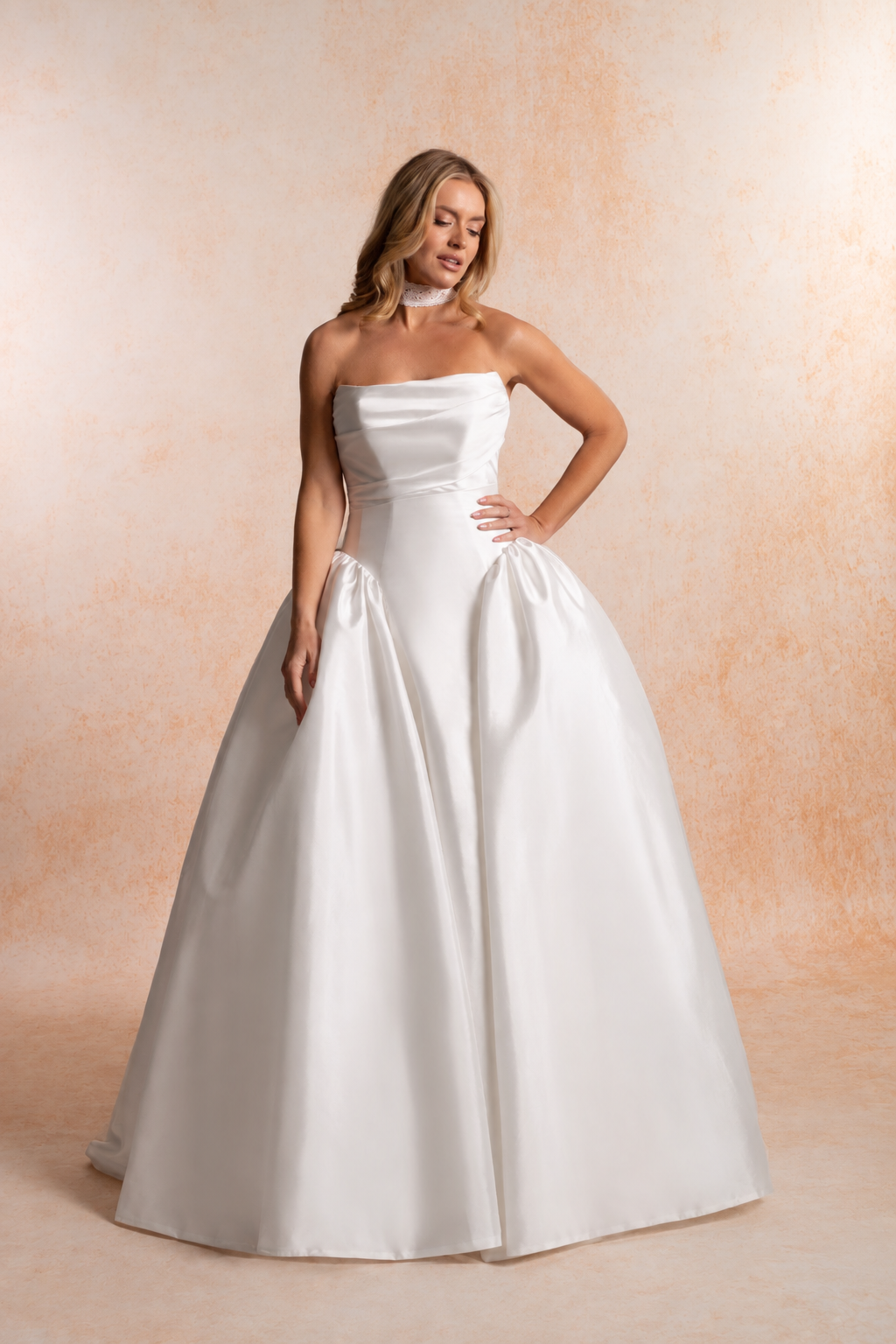 Model wearing a strapless taffeta ball gown wedding dress with a straight neckline, softly draped bodice, elongated pointed waist and full gathered skirt.