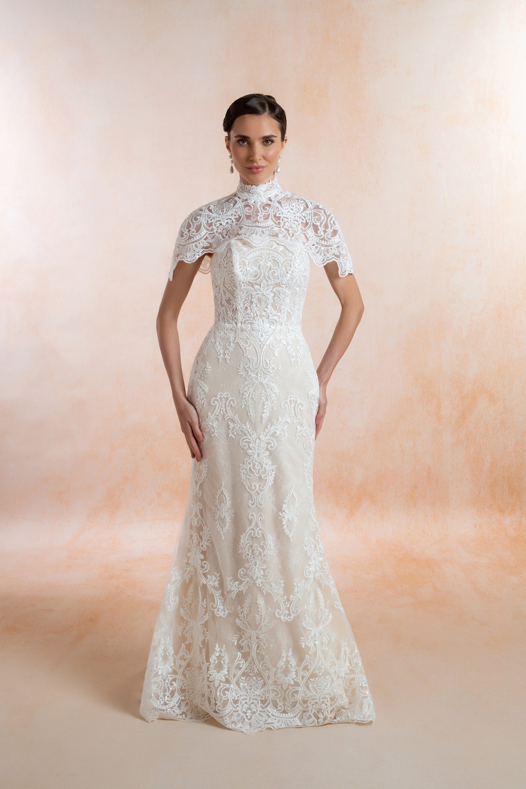 Model wearing an ivory lace fit-and-flare wedding dress styled with a high-neck lace cape, with a defined waist and gently flared skirt.